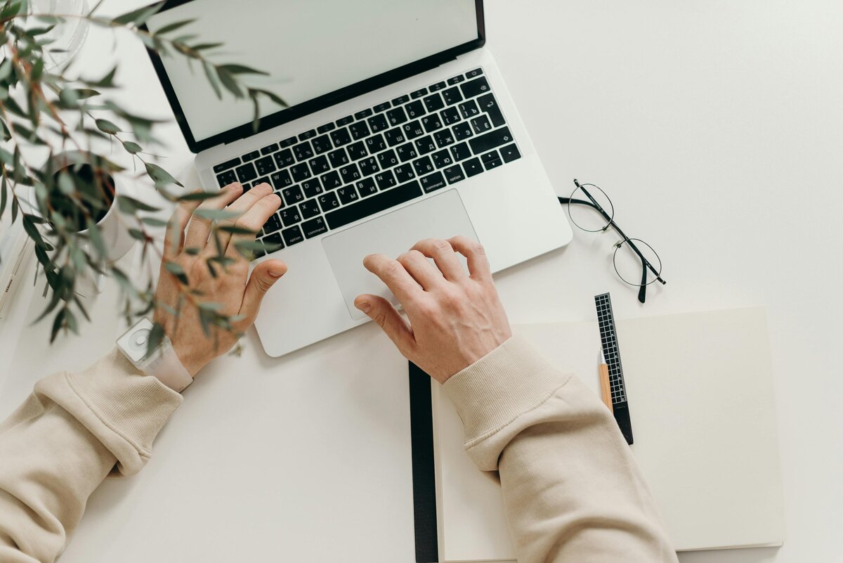 A person wearing a beige sweatshirt working on a laptop at a minimalist white desk. Surrounding the laptop are a pair of glasses, an open notebook with a pen, and a potted plant partially covering the frame.
