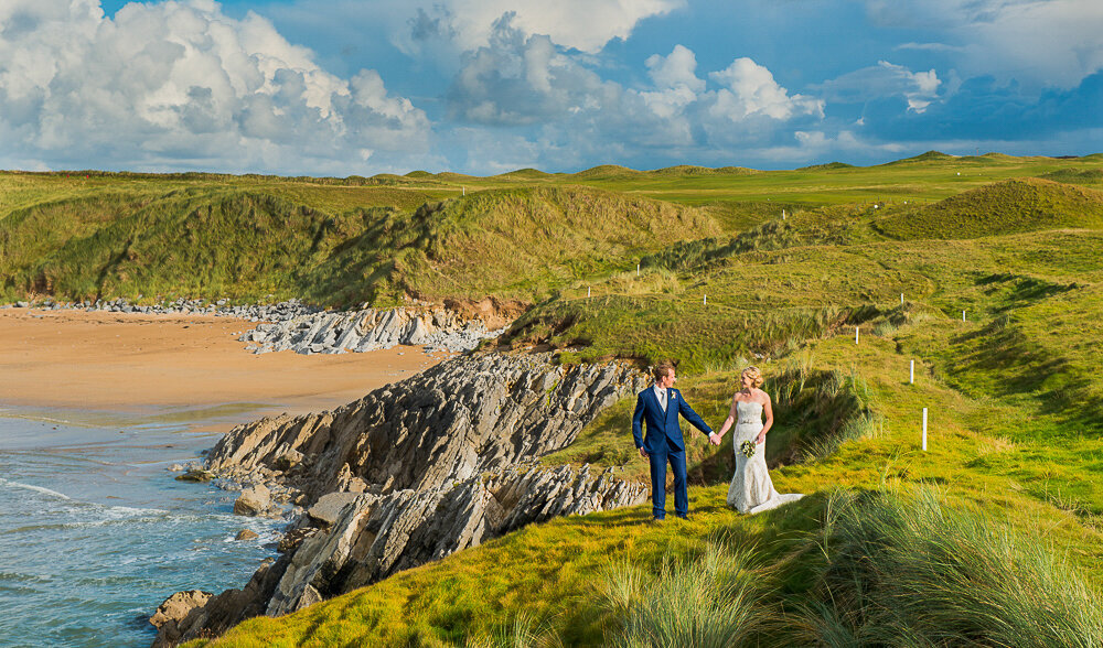 Bride in A-line wedding dress holding hands with red haired groom wearing a navy suit, standing on cliffs overlooking the beach in Kerry