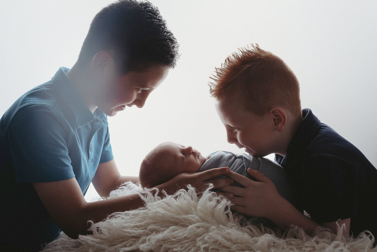 Newborn baby laying on white fur with big brothers kneeling down and leaning in looking at baby.
