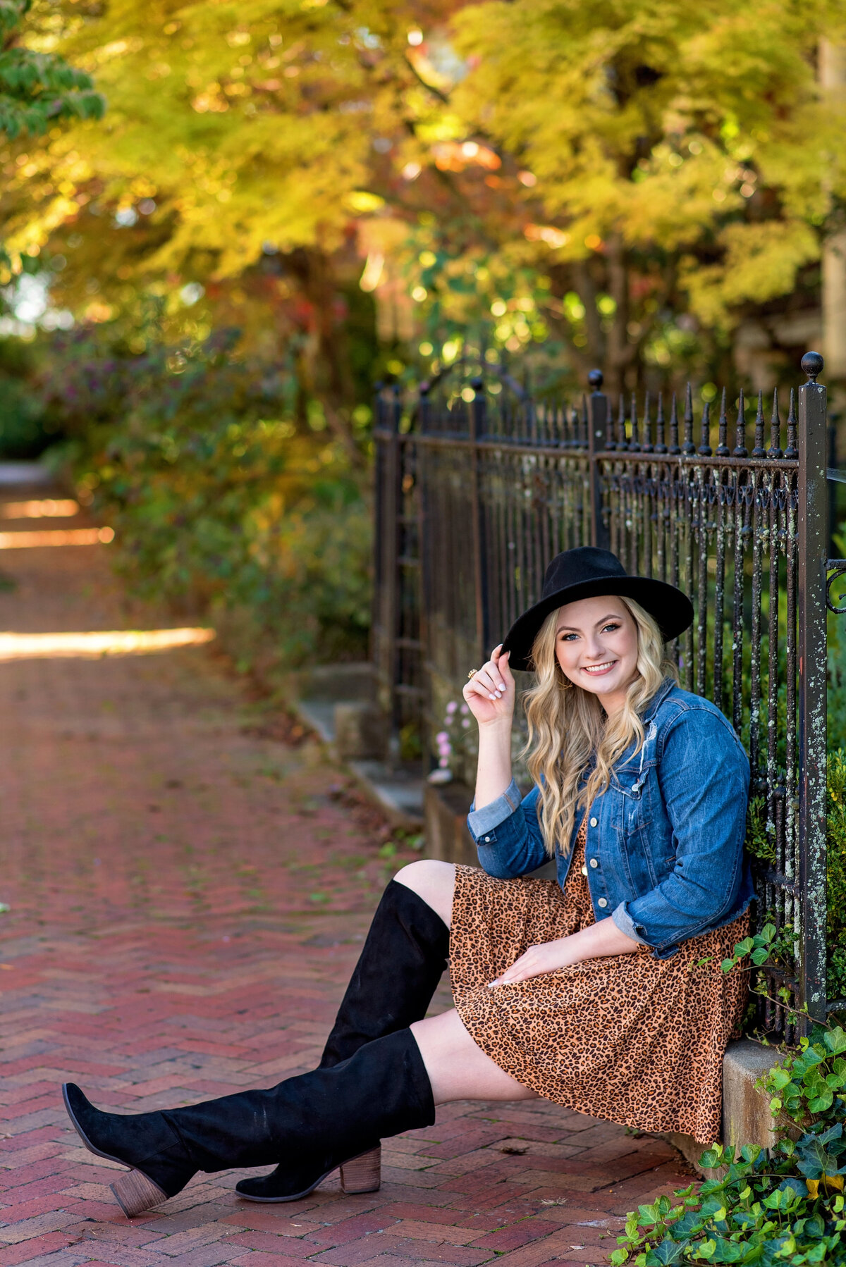 Richmond high school senior girl sits against iron fence in Libby Hill during fall wearing denim jacket, black boots, and black hat for her senior portraits.