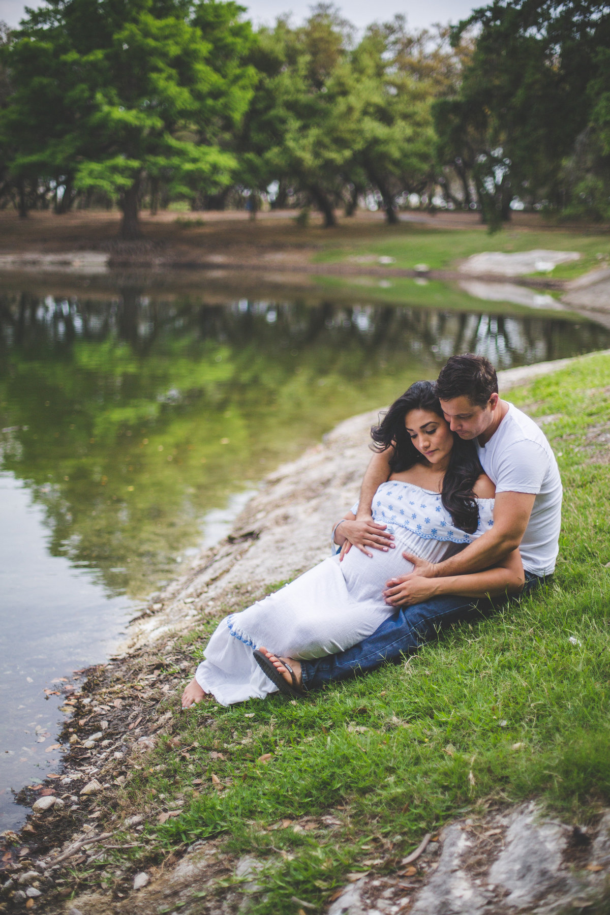 Man holding his pregnant wife wearing a maternity dress while sitting by a lake at Denman Estate park in San Antonio.
