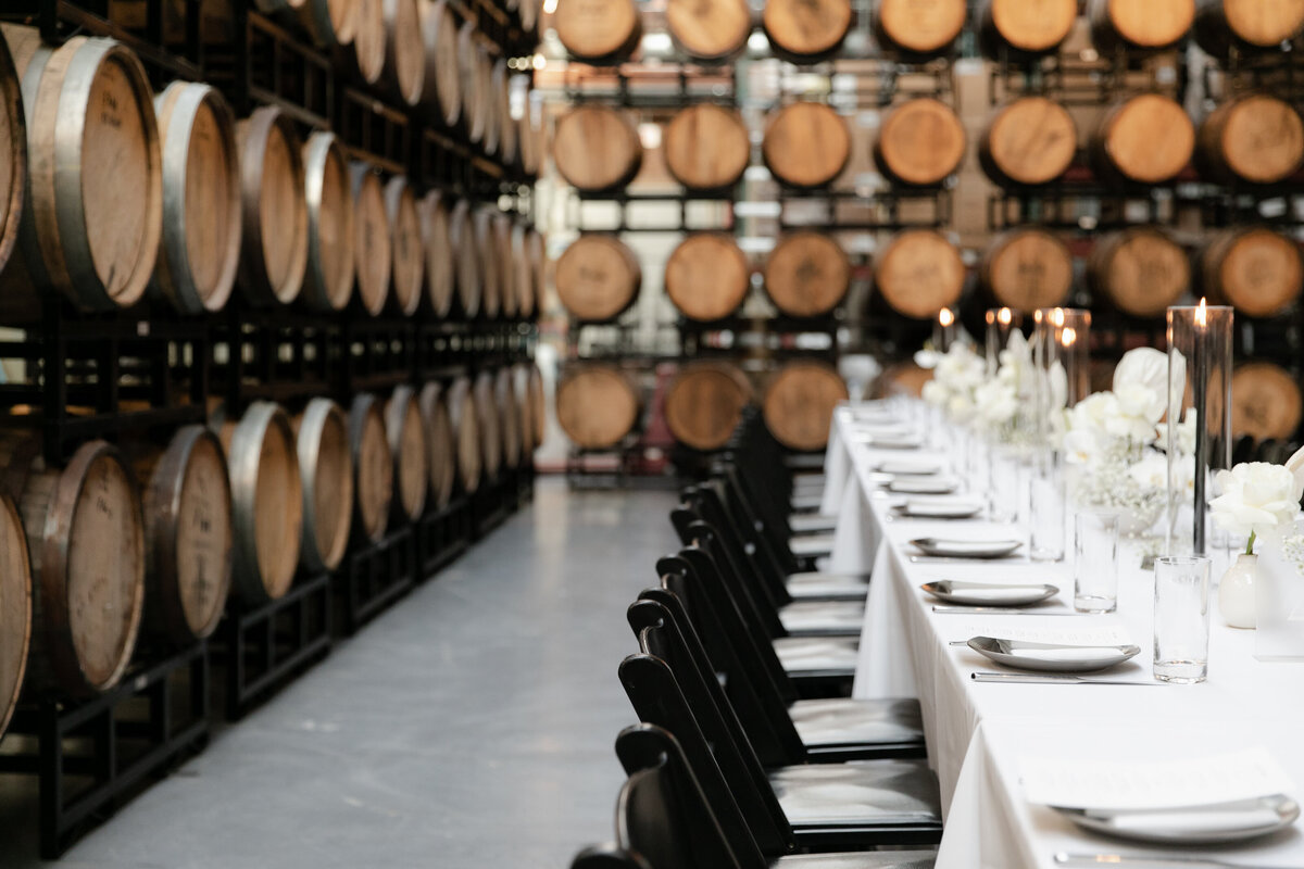 Simple centerpieces composed of black candlesticks, glassware and white lilies sit on long wedding reception table lined with black chairs in Mood Tongue Brewery with beer barrels pictured in the background.