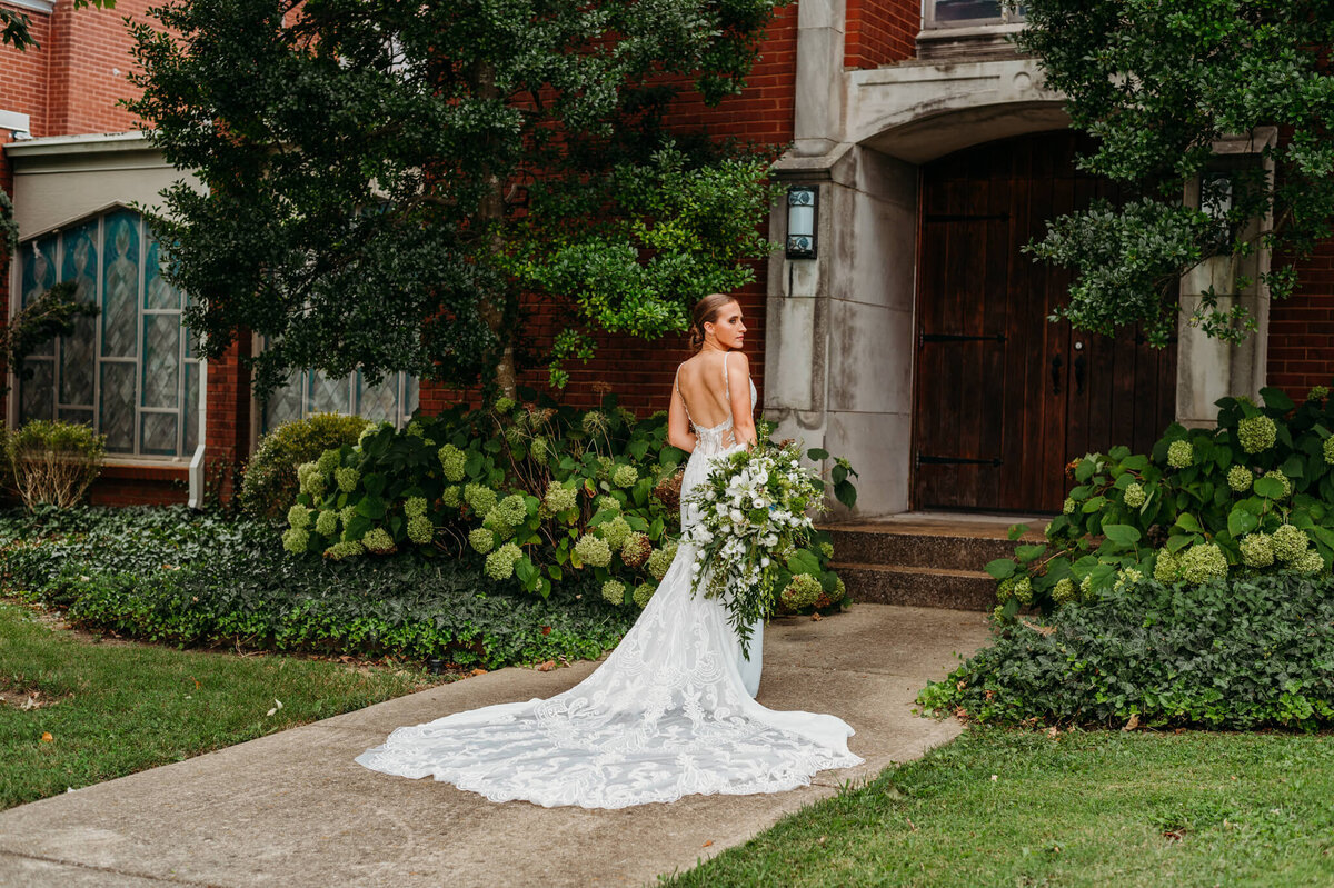 Photo of the back of a brides dress well she looks to the side in front of a church with hydrangea bushes and a wooden door