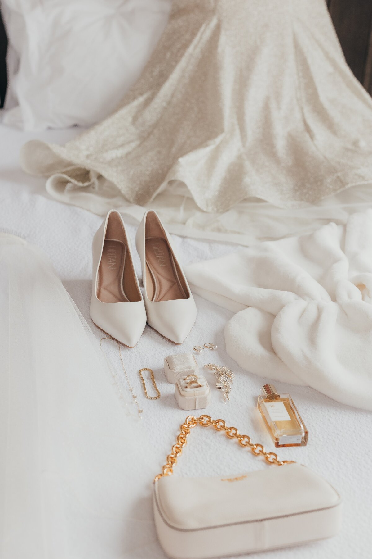 A pair of white high-heeled shoes, jewelry, and a purse arranged on a soft fabric surface near a flowing white dress, ideal for Iowa weddings.