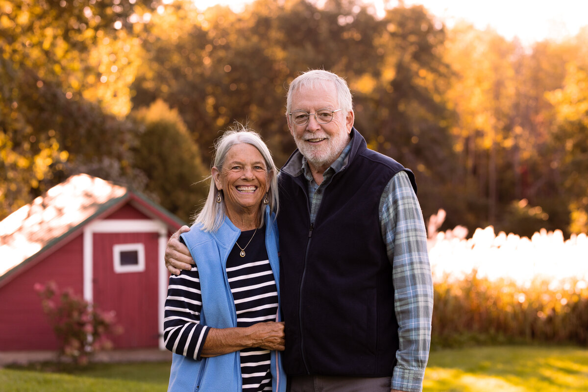 Portrait of grandparents in front of a red sheep shed in the fall by  Vermont Family Photographer