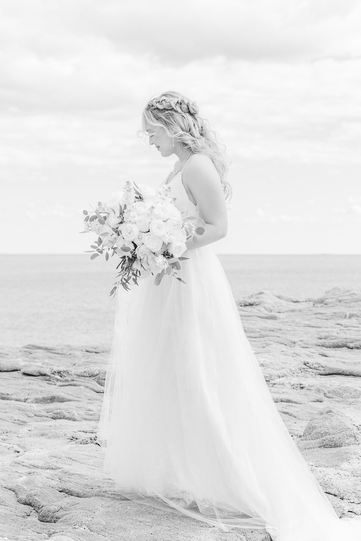 Bride is standing on the rocks along the shoreline at the Madison Beach Hotel for a candid image. She is standing side profile to the camera and looking down as she walks. Captured by Connecticut wedding photographer Lia Rose Weddings