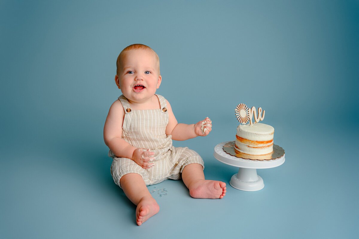 Simple smash themed setup with a little boy dressed in a cream colored romper sitting on a blue background with his cake beside him with a cute little cake topper