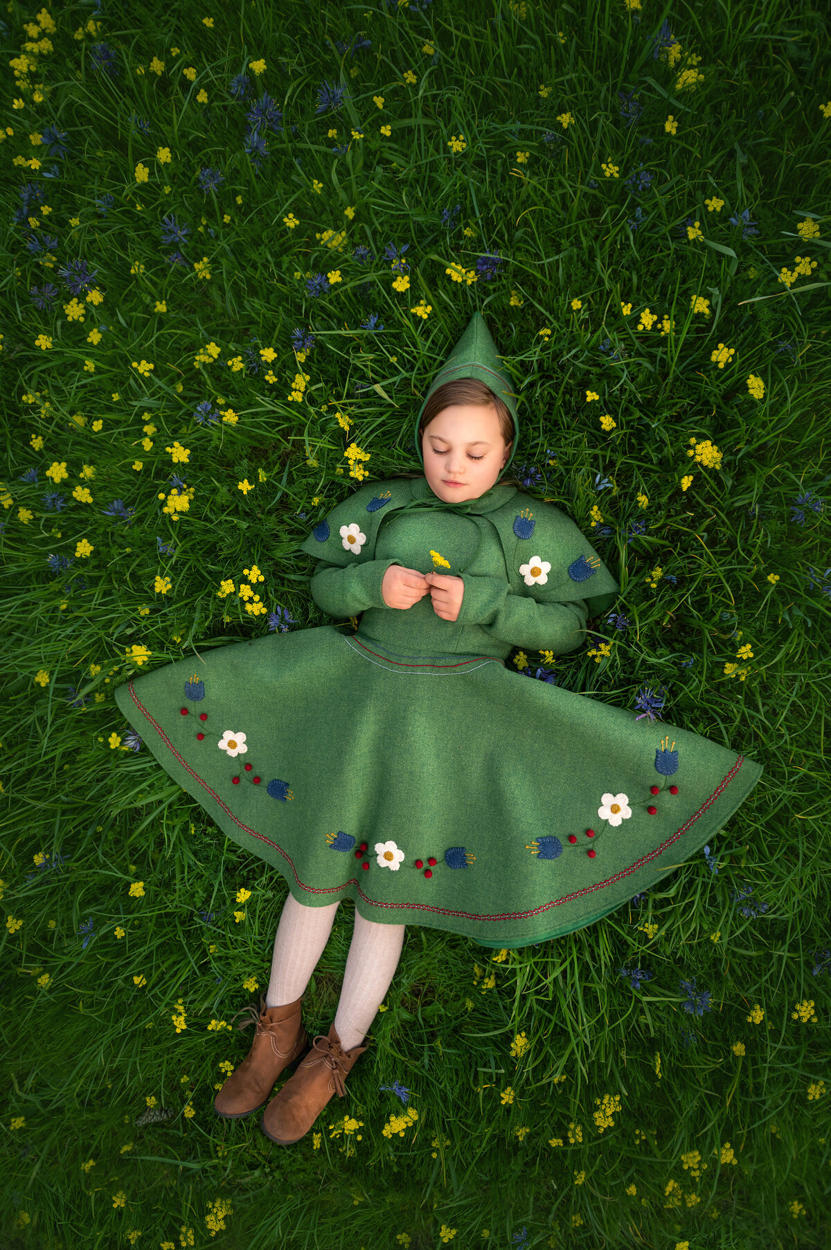 A girl lays flat on her back in a field of yellow flowers wearing gnome-inspired costuming.