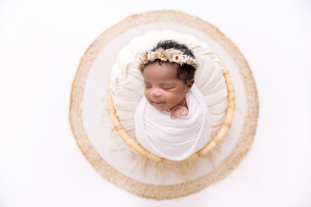 A newborn baby girl sleeps in a white swaddle and floral headband inside a wicker basket in a studio