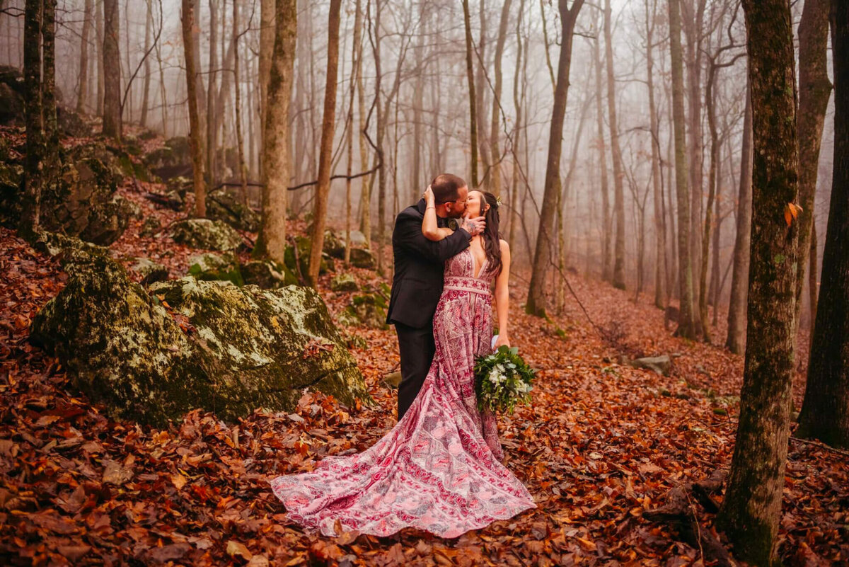 Photo of a bride and a purple gown kissing her groom in the forest