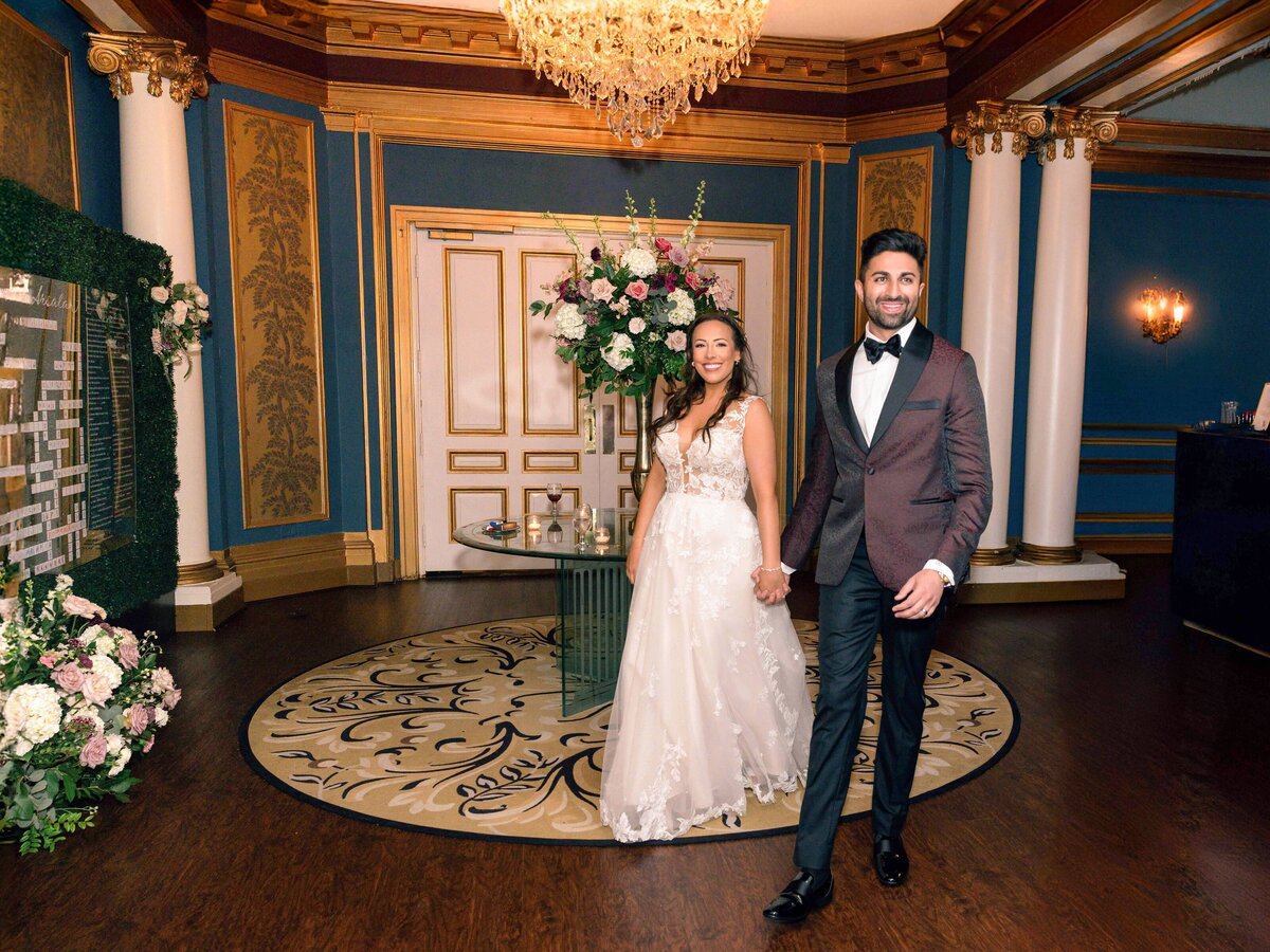 A couple in formal attire walks hand in hand in an elegant room with ornate blue and gold walls, a chandelier, and a large floral arrangement. The woman wears a white gown, and the man wears a suit with a bow tie.