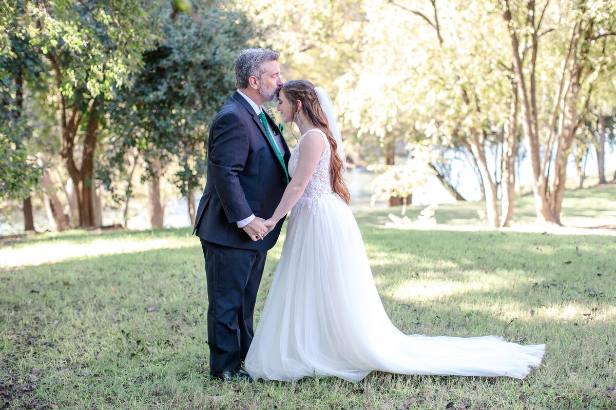 father kisses bride's forehead while holding her hands under the trees at Milltown Historic District wedding in New Braunfels