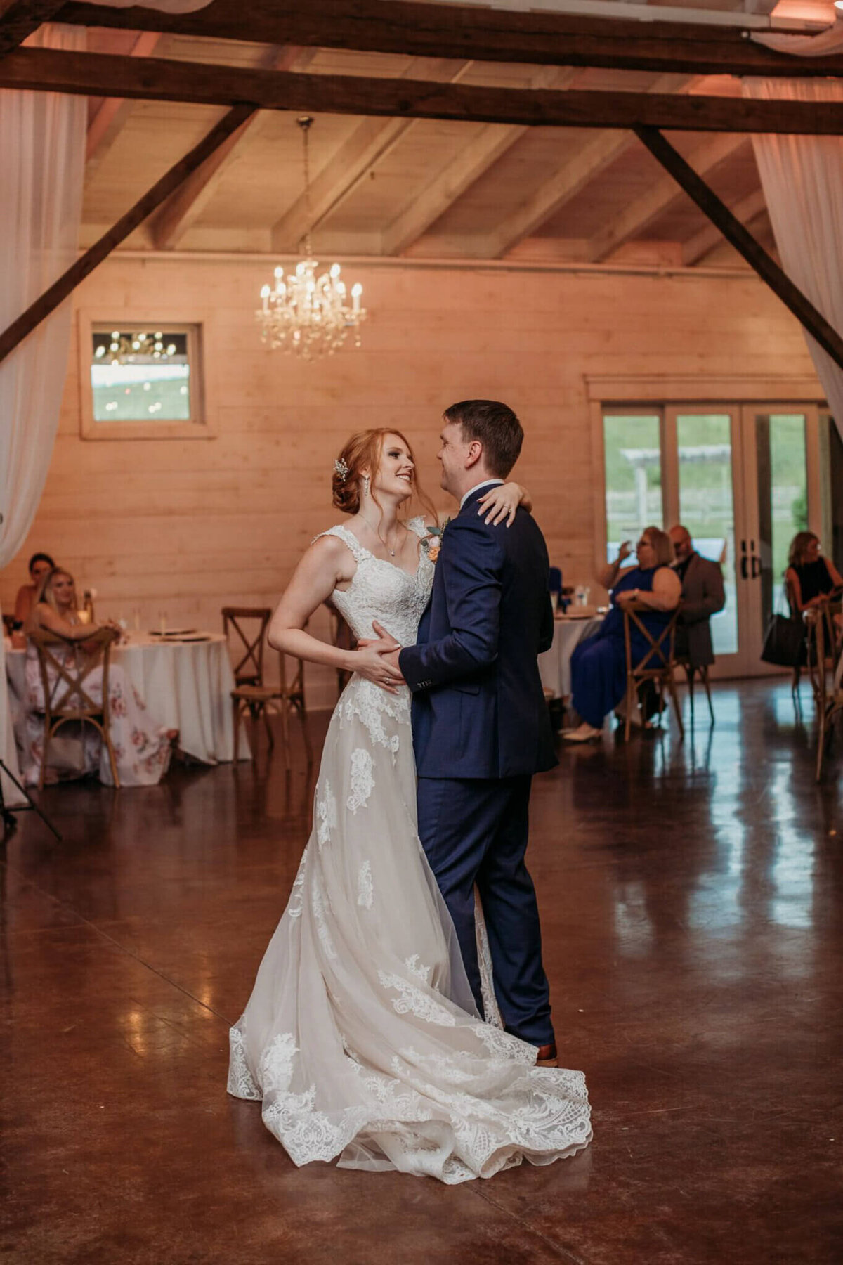 Photo of a bride and groom slow dancing inside of a white washed barn with chandeliers