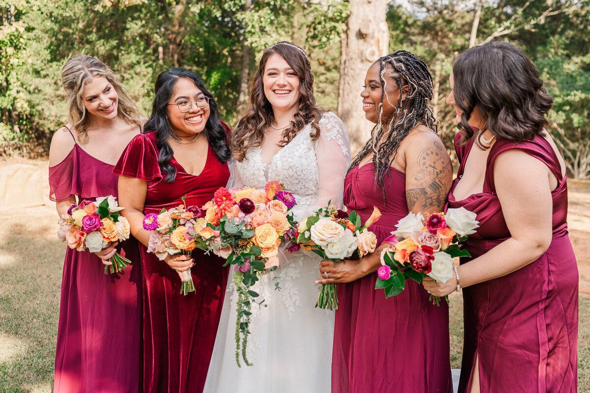 A laughing bride surrounded by her six bridesmaids in maroon dresses during her fall wedding enjoying her Raleigh wedding photography session with JoLynn Photography