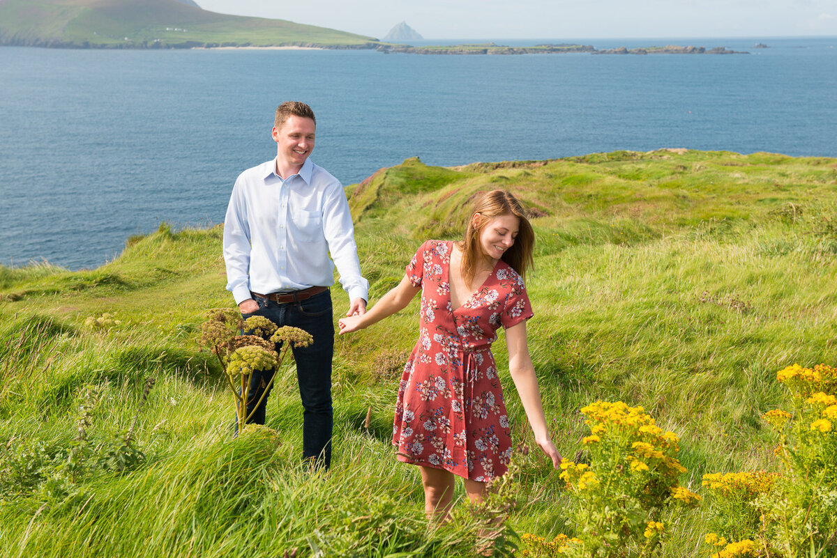 Young couple holding hands and standing in a green field with yellow flowers and overlooking the sea