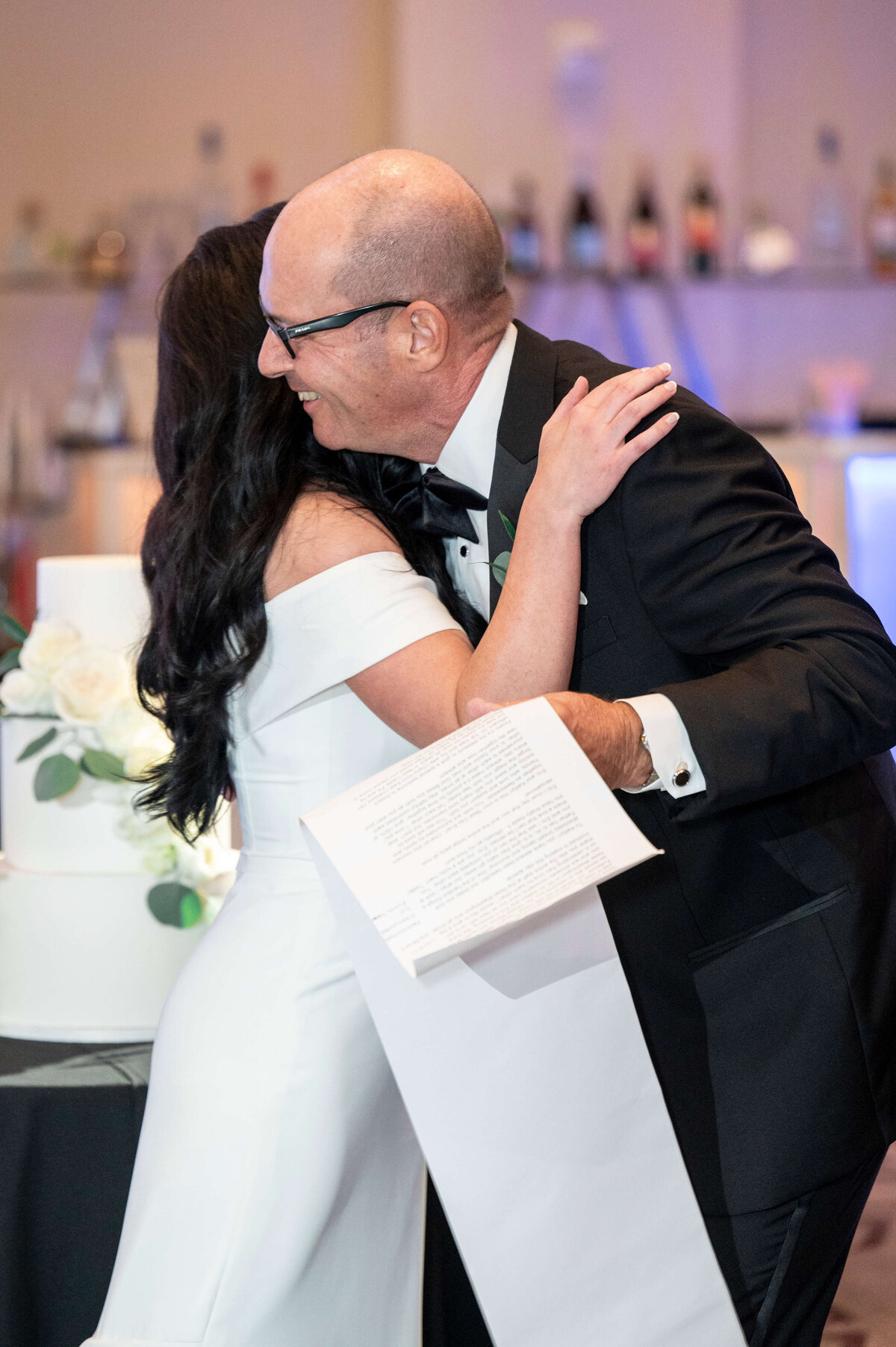 A man in a tuxedo hugs a woman in a white dress at a formal event. He holds papers in one hand, possibly wedding vows or a speech. A decorated cake is visible in the background, suggesting a celebration or wedding reception.