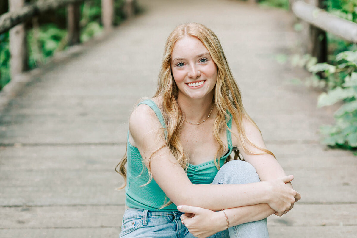 A high school senior sits on the bridge at Beazell Memorial Forest for Senior portraits. She's wearing a green tank top and jeans and has her knee up and arms wrapped around the knee
