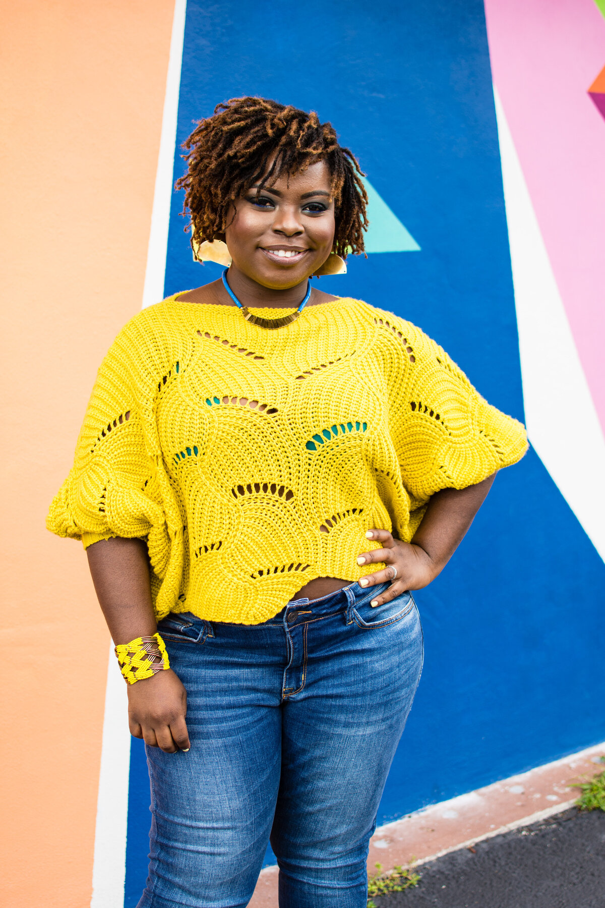 Annie Brown poses in a yellow crochet top and jeans in front of a colorful mural