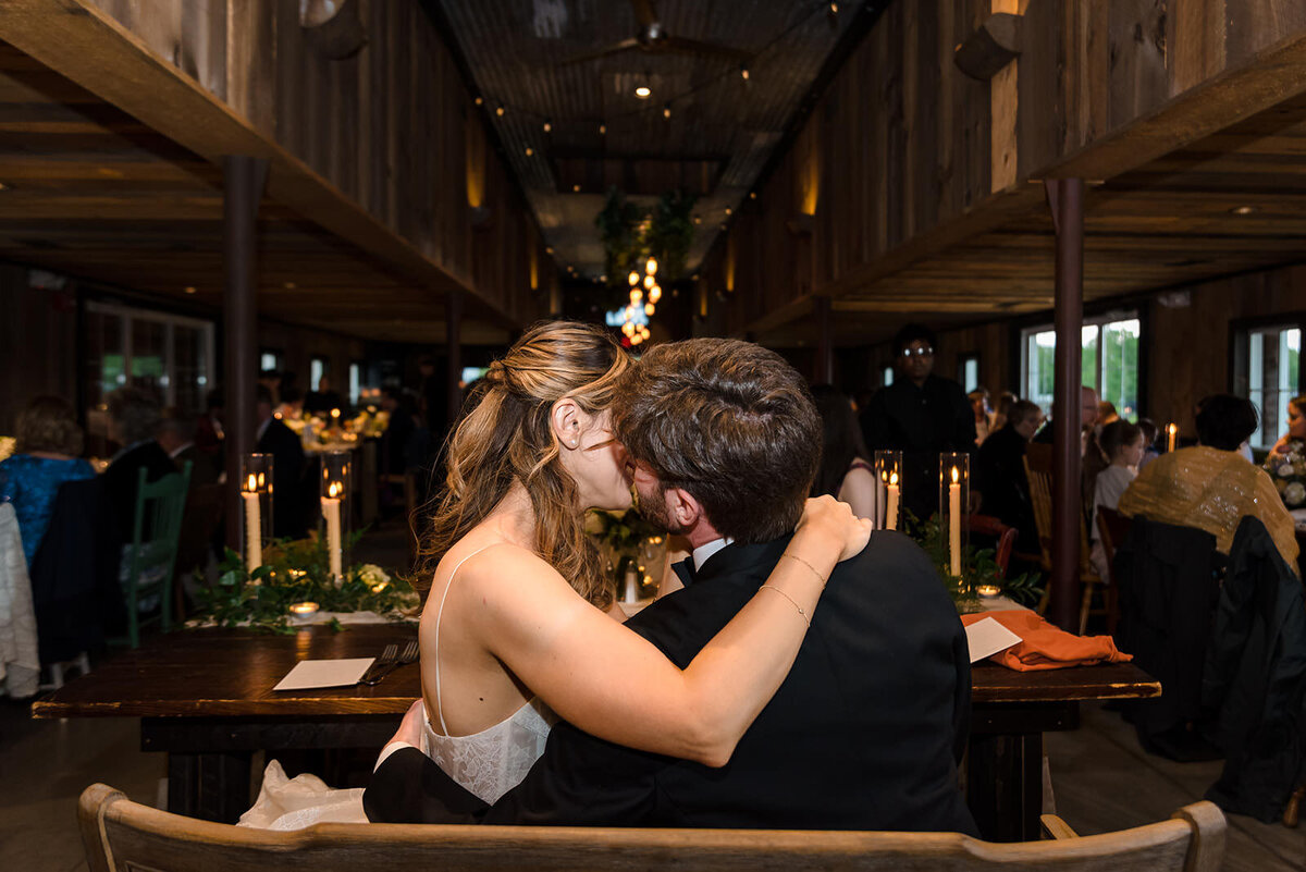 A bride and groom kissing at their wedding reception, with guests and the lit-up barn interior in the background.
