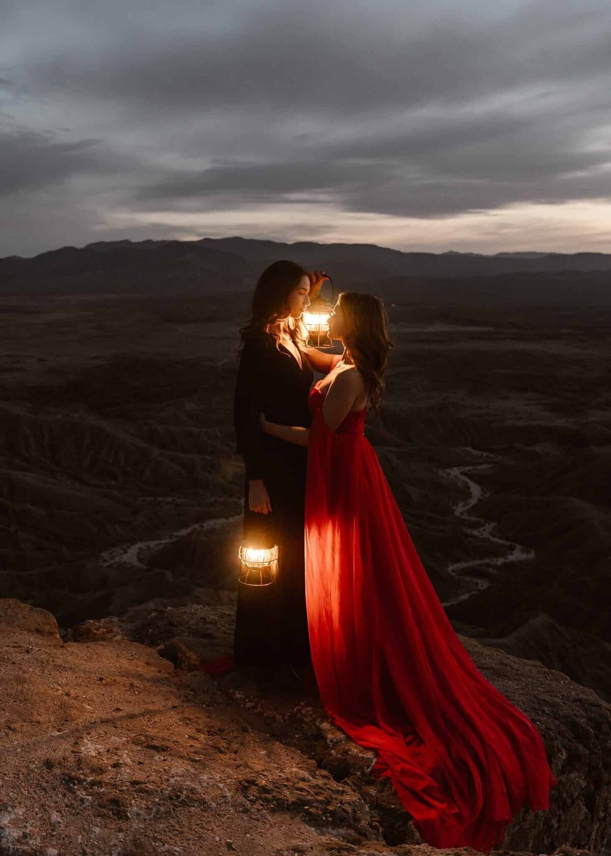 A couple holding lanterns stands close together on a cliff at dusk, one in a red dress and the other in black, with soft light from the lanterns lighting their faces.