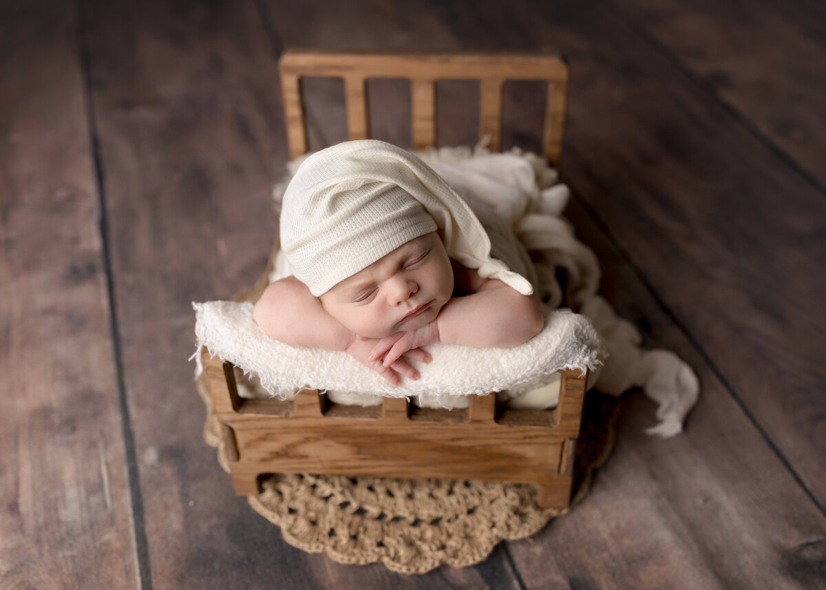 Baby boy newborn photoshoot. Baby is sleeping on a miniature bed with his hands folded under his chin and his cheek is resting on his left arm. Baby is wearing a long cream cap with the end trailing over his shoulder.