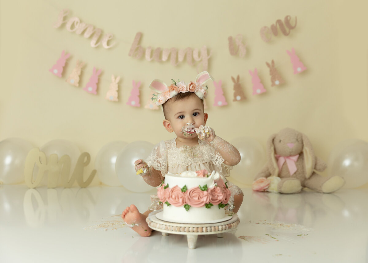 A toddler girl sits in a studio wearing a beige dress eating a cake