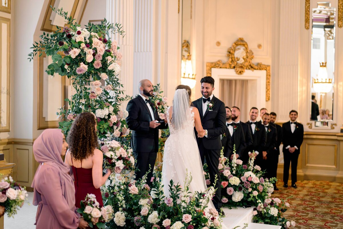 A wedding ceremony takes place in an ornate room with floral decorations. The couple stands under a floral arch facing the officiant. Bridesmaids and groomsmen are present, with guests seated nearby. The setting is elegant and formal.