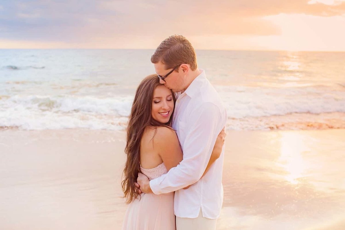 Man wearing glasses lovingly kisses his wife on the forehead as she smiles and looks down. There is a beautiful Maui sunset behind them.