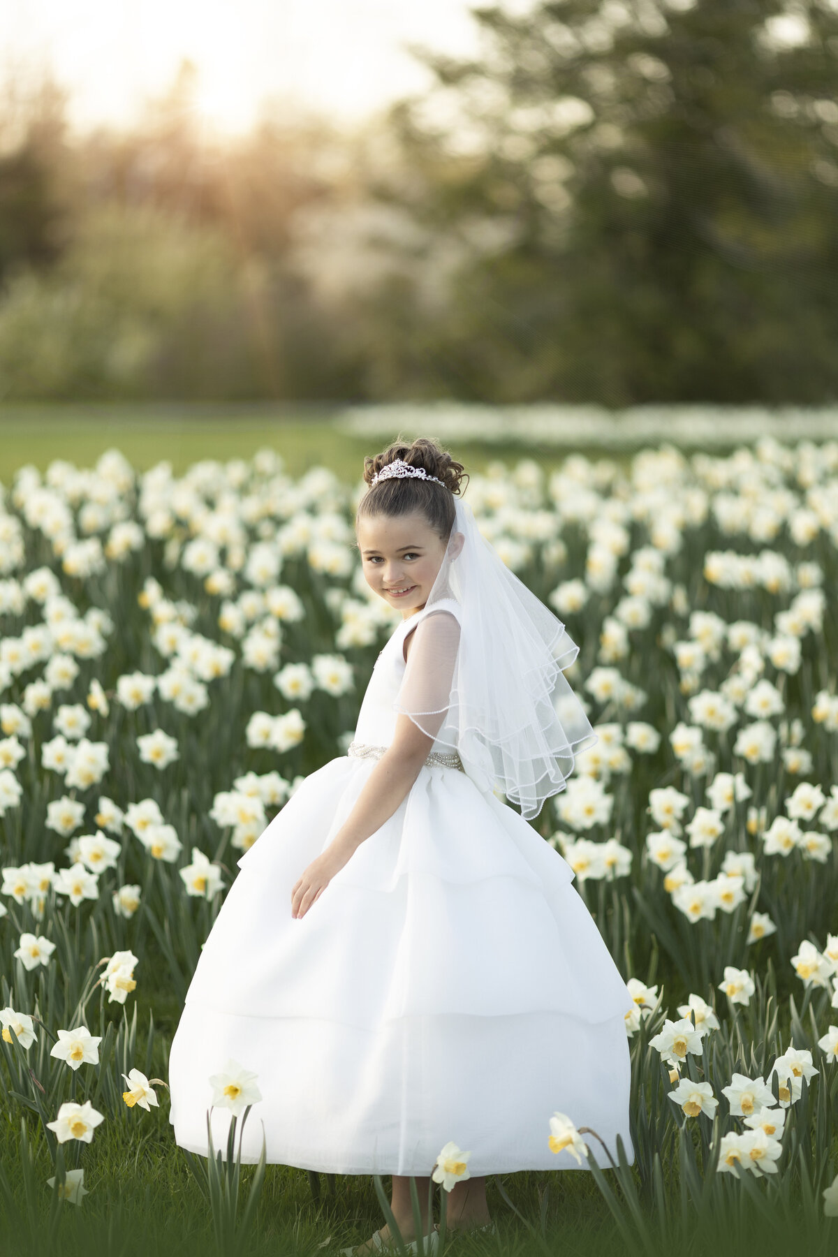 A girl in a white poofy dress walks through a field of white daffodils at sunset
