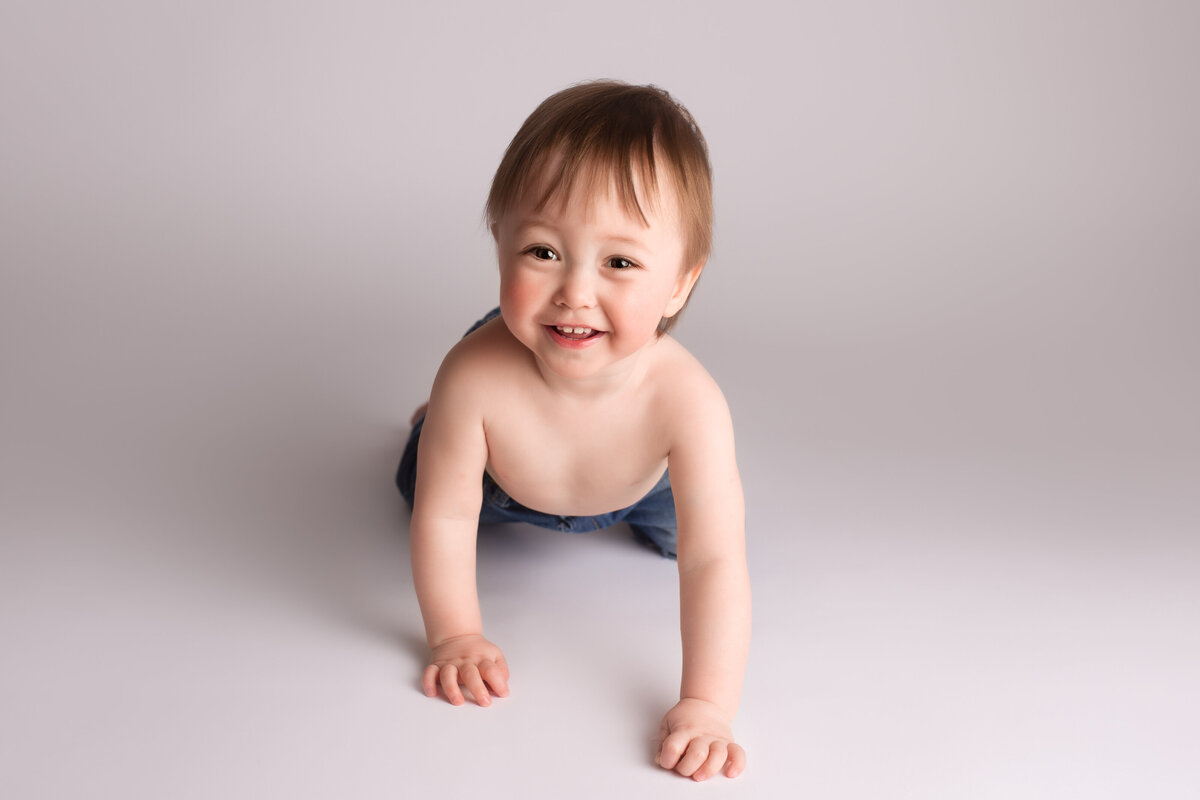 Baby in a crawling position wearing jeans looking up and smiling on a simple white background.