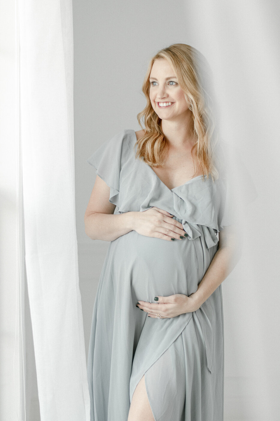 Pregnant woman in a. blue dress smiles out the window By Nashville maternity photographer Kristie Lloyd