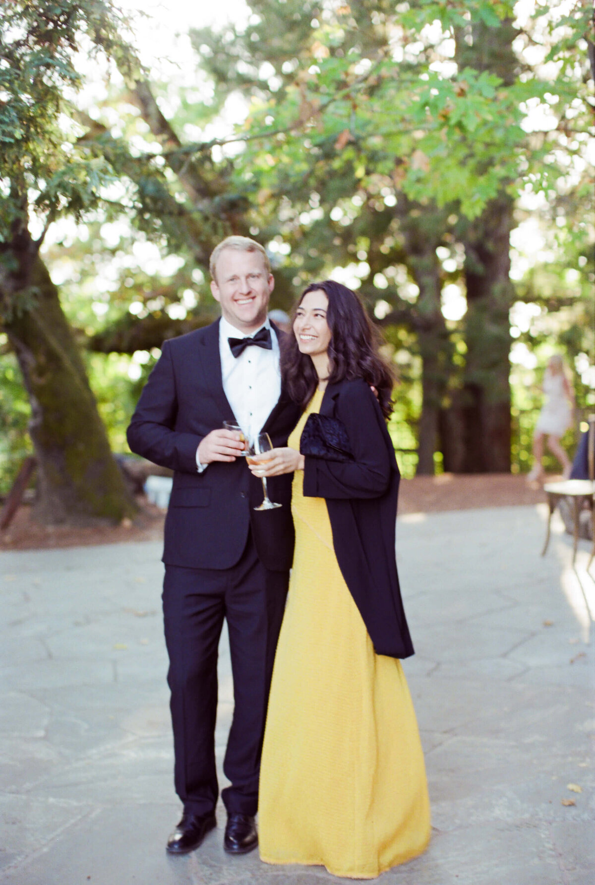 Man in black suit and bow tie with a lady in lemon empire waist gown pose with champagne at a vineyard wedding venue.
