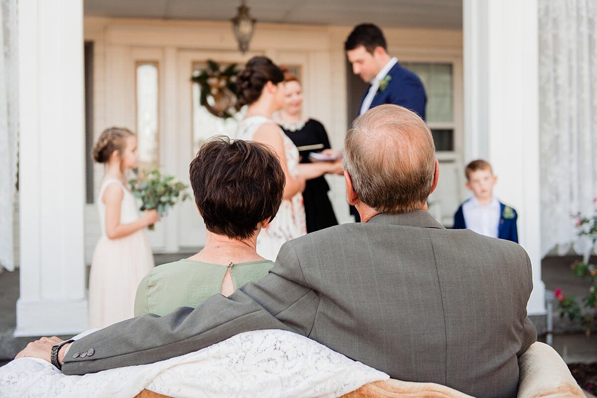The bride's parents watch the wedding ceremony from their seats. The bride, groom, officiant, flower girl and ring bearer are all standing on the front porch of a large white mansion.