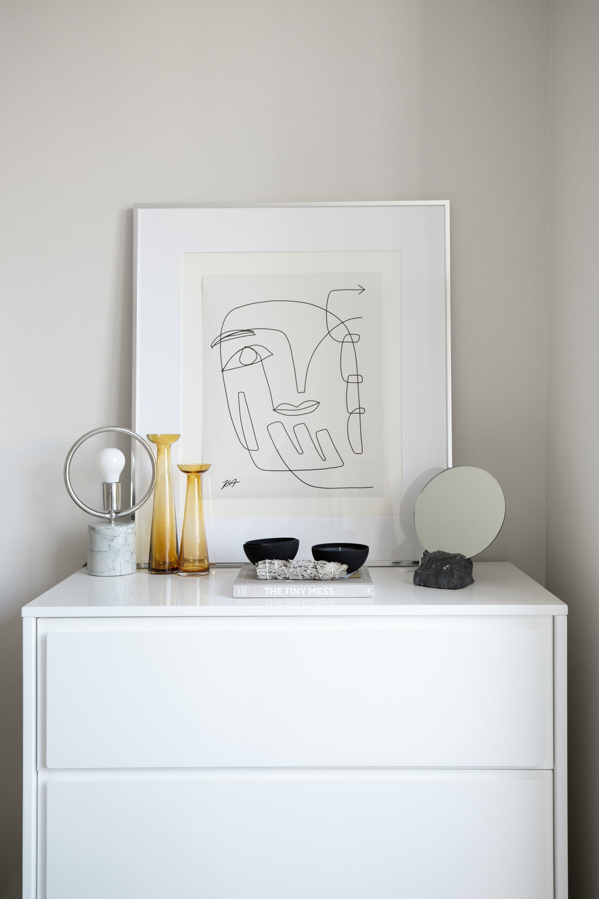 Lacquered white dresser with chrome framed abstract portrait artwork, round vanity mirror, yellow glass vases, candle, sage and circular chrome marble table lamp