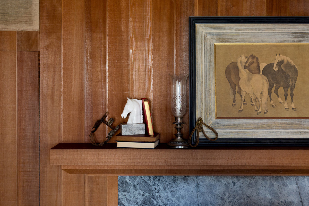 Redwood mantel with books, western horse bit, white marble horse head bookend, cut glass hurricane, and framed print of oriental horses