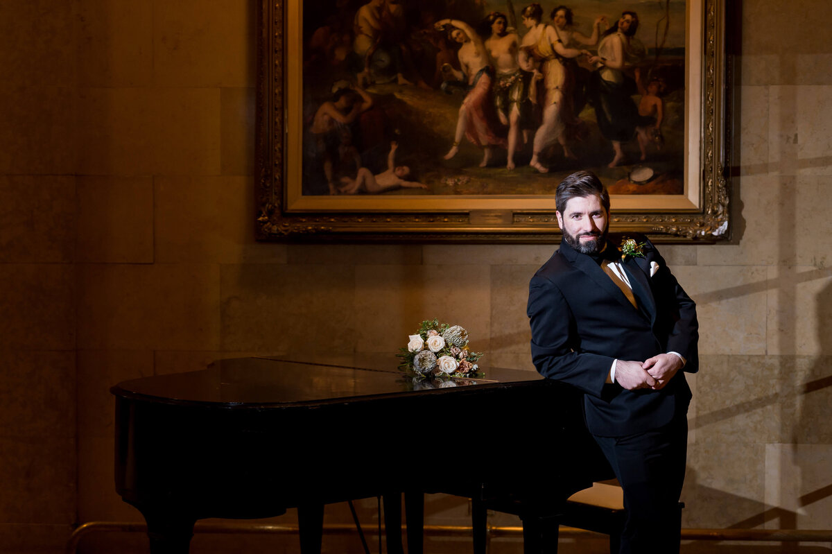 a groom in a black tuxedo leans on the grand piano of the Chateau Laurier hotel.  Photographed by Ottawa wedding photographer JEMMAN Photography