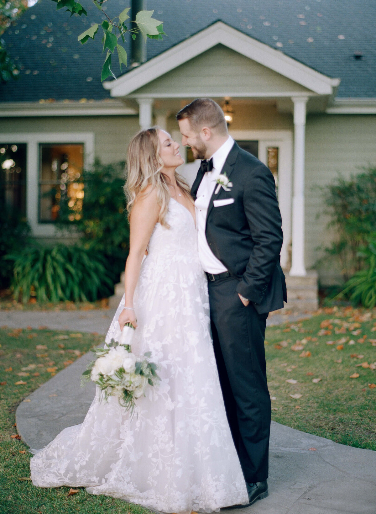 Just-married bride and groom share an intimate moment together in front of an American-style country home. Dressed in sophisticated Christian wedding attires, the wife holds a white bouquet while a maple tree branch hangs from top.