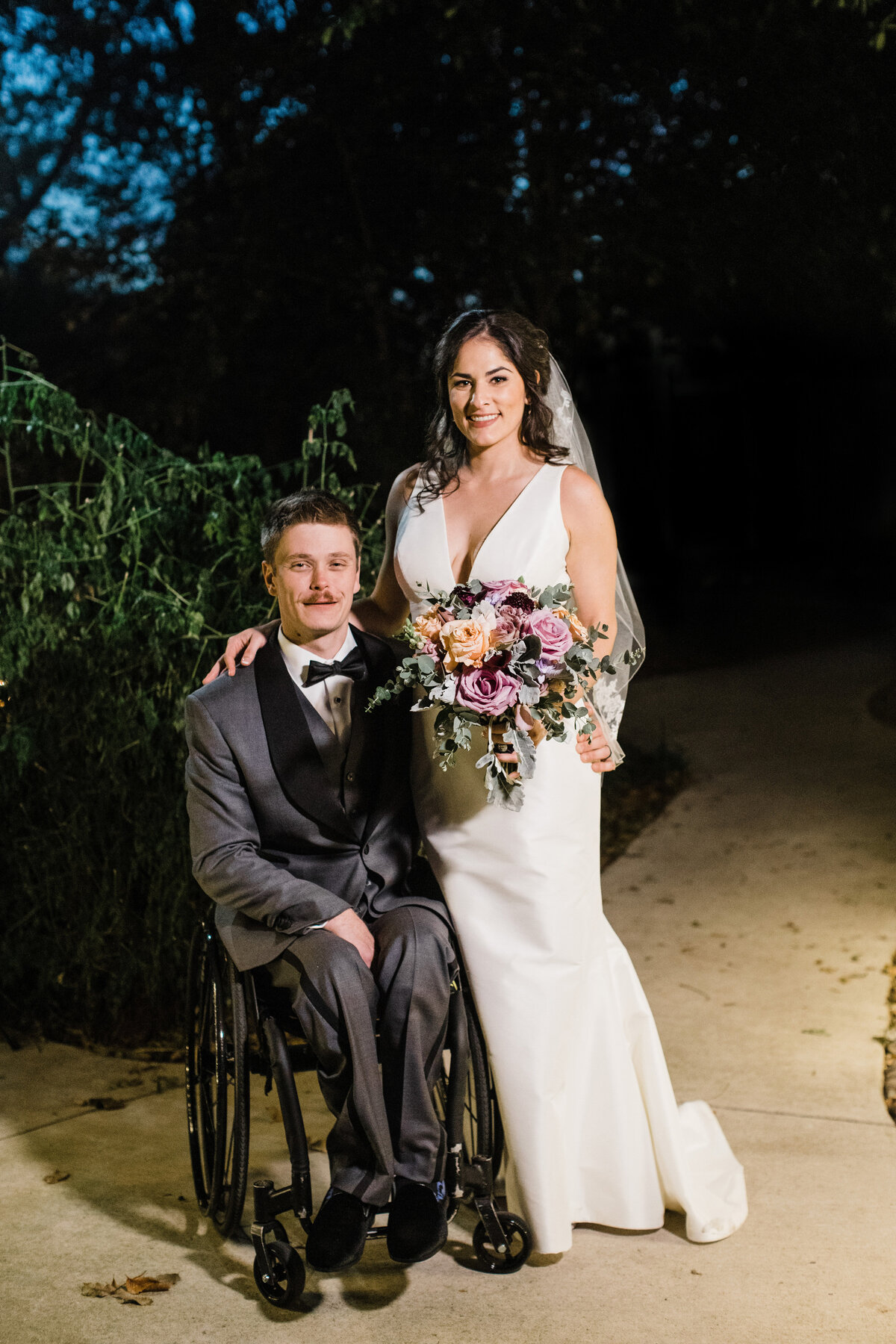 A nighttime portrait of a bride and groom after their wedding ceremony in DFW, Texas. The bride is on the right and is wearing a sleeveless, white dress with a veil and is holding a bouquet. The groom is on the right and sits in a wheelchair while wearing a grey suit and bowtie.