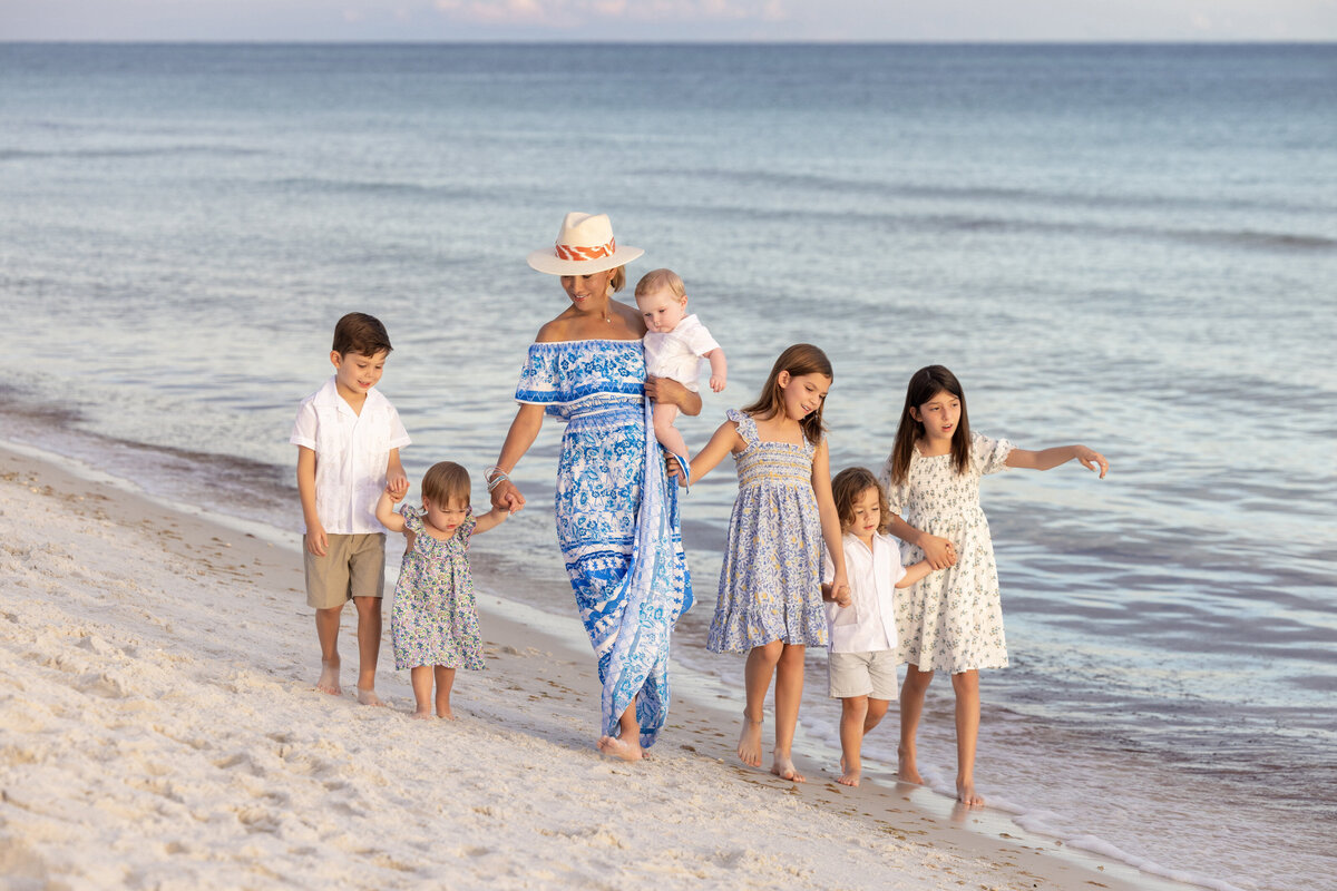 A woman walking along a beach holding a child and walking with five other kids