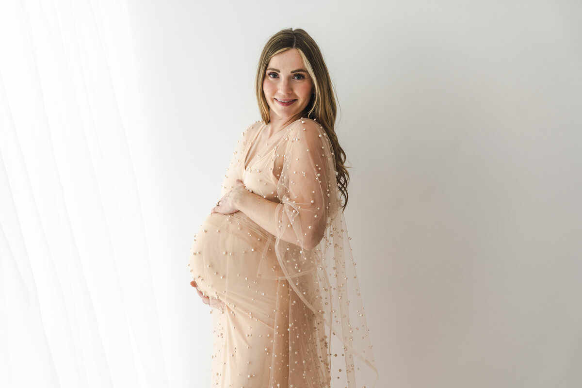 A radiant expectant mother in a beautiful beige dress adorned with sparkles, cradling her baby bump with a joyful smile against a minimalist white background. Taken by Minneapolis maternity photographer, Fig and Olive Photography.
