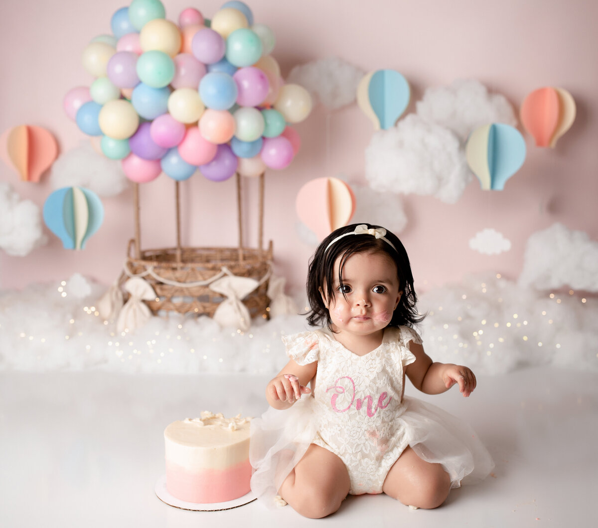 Girly hot air balloon themed cake smash in West Palm Beach, FL newborn and cake smash photography studio.  Baby girl is sitting on her knees in a white dress looking at the camera with a pink ombre cake beside her. in the background, there are pastel-colored hot air balloons, faux clouds and twinkle lights.