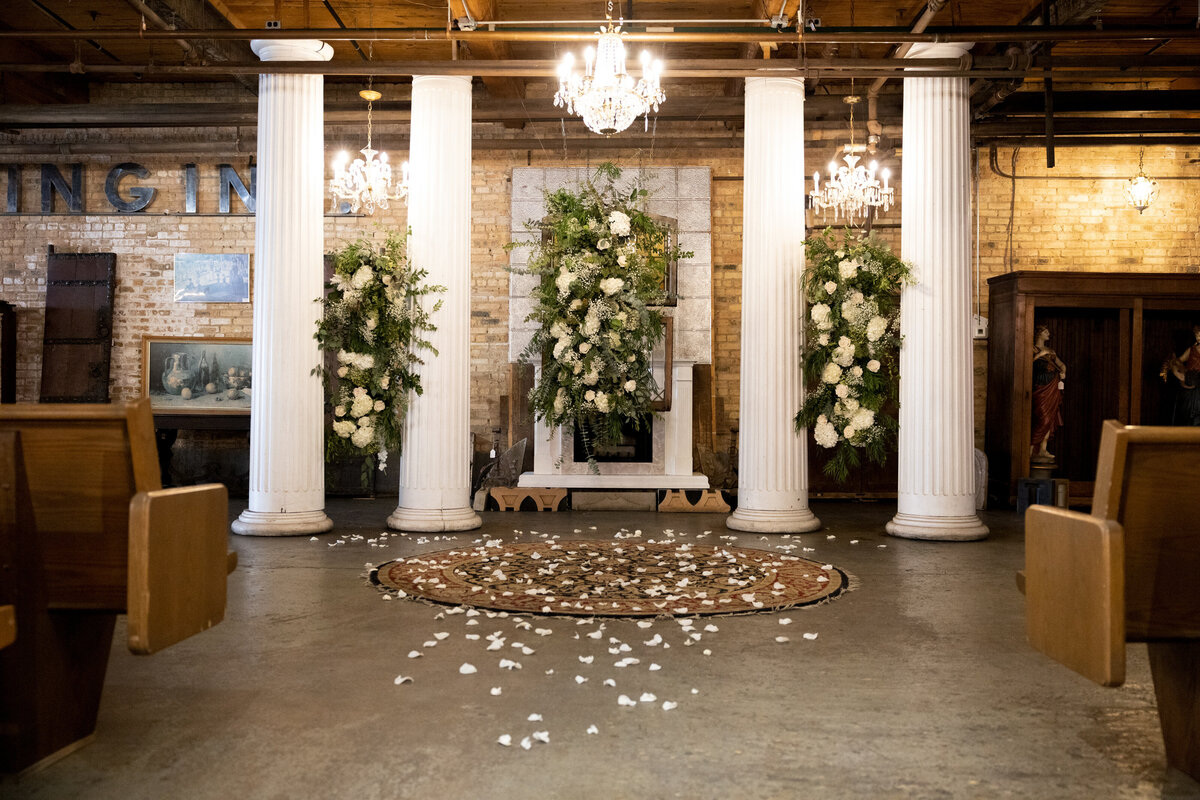 White rose pedals are spread on the floor near wedding alter comprised of four white columns, draping greenery and white flowers and chandeliers.