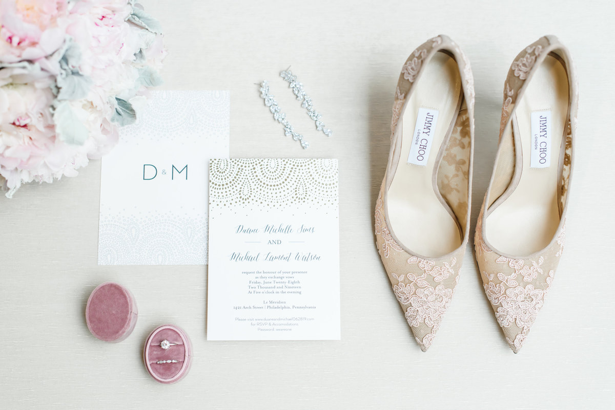 bridal details consisting of a pink and white bouquet, ivory and gold invitation, rose colored ring box holding bride's rings and lace jimmy choo high heels