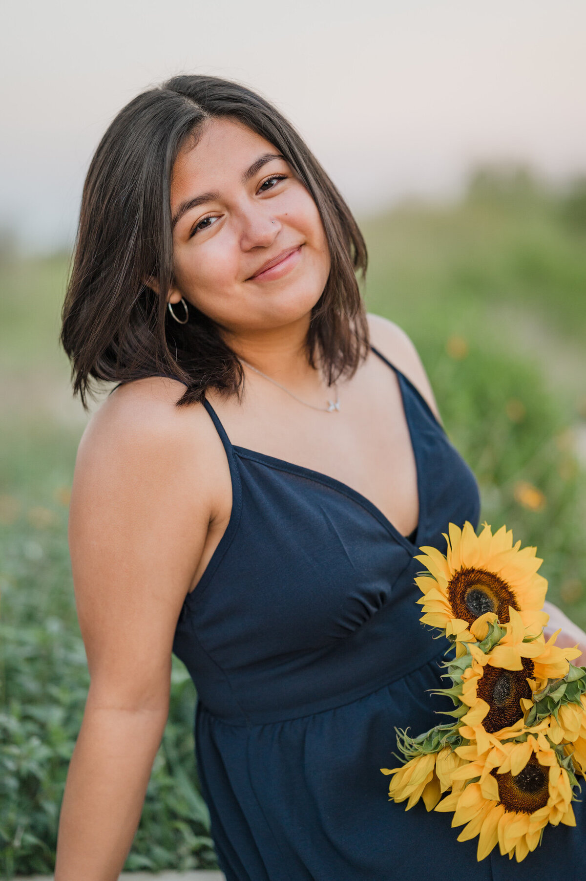 Girl in a blue sundress holds sunflowers and smiles for senior portraits in San Antonio.