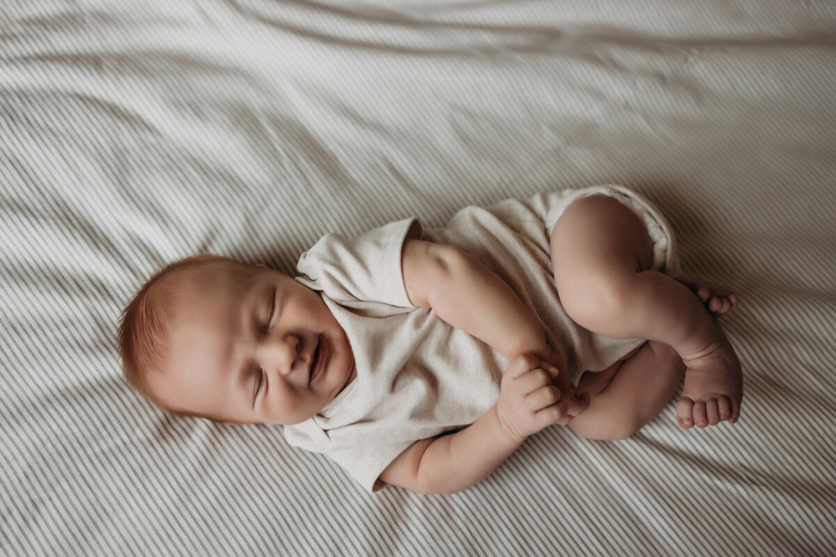 overhead photo of baby boy scrunched up in a white onesie on bed