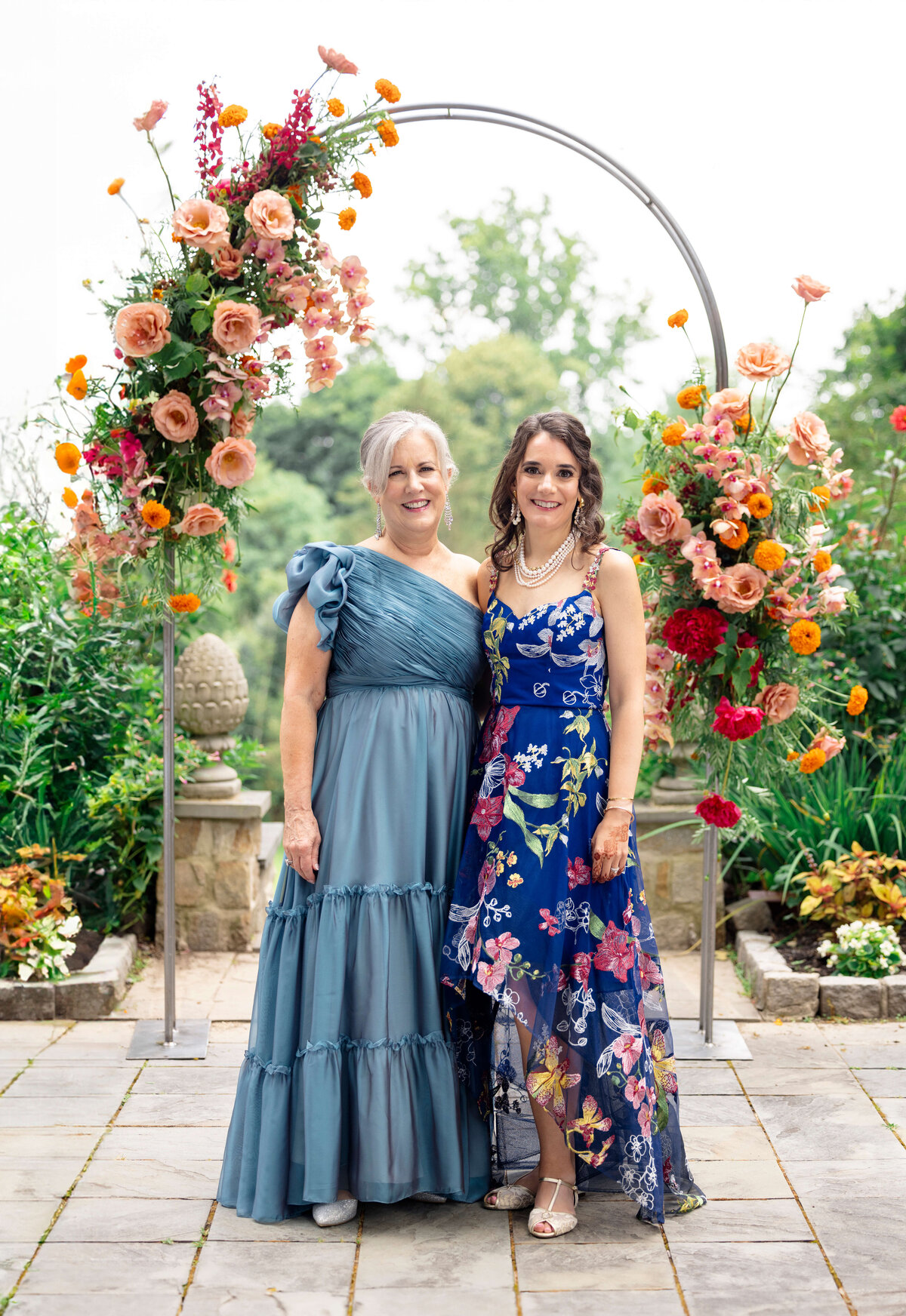Two women stand smiling under a circular floral arch. One wears a blue ruffled gown, and the other wears a blue floral dress. The background features lush greenery and colorful flowers.