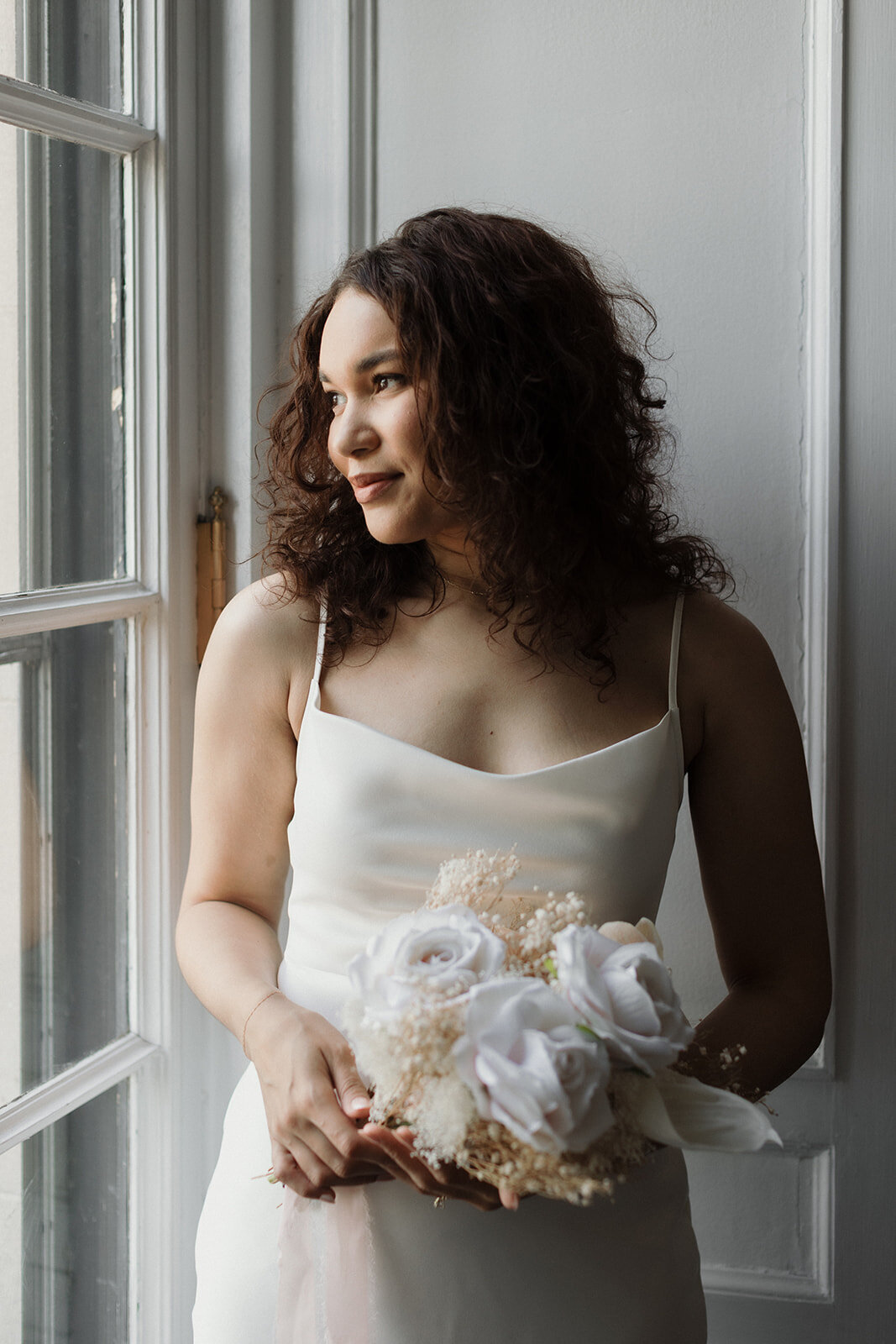 Just Married photo session bride stands near window with beautiful ight and looks out the window. She holds her bouquet gently.