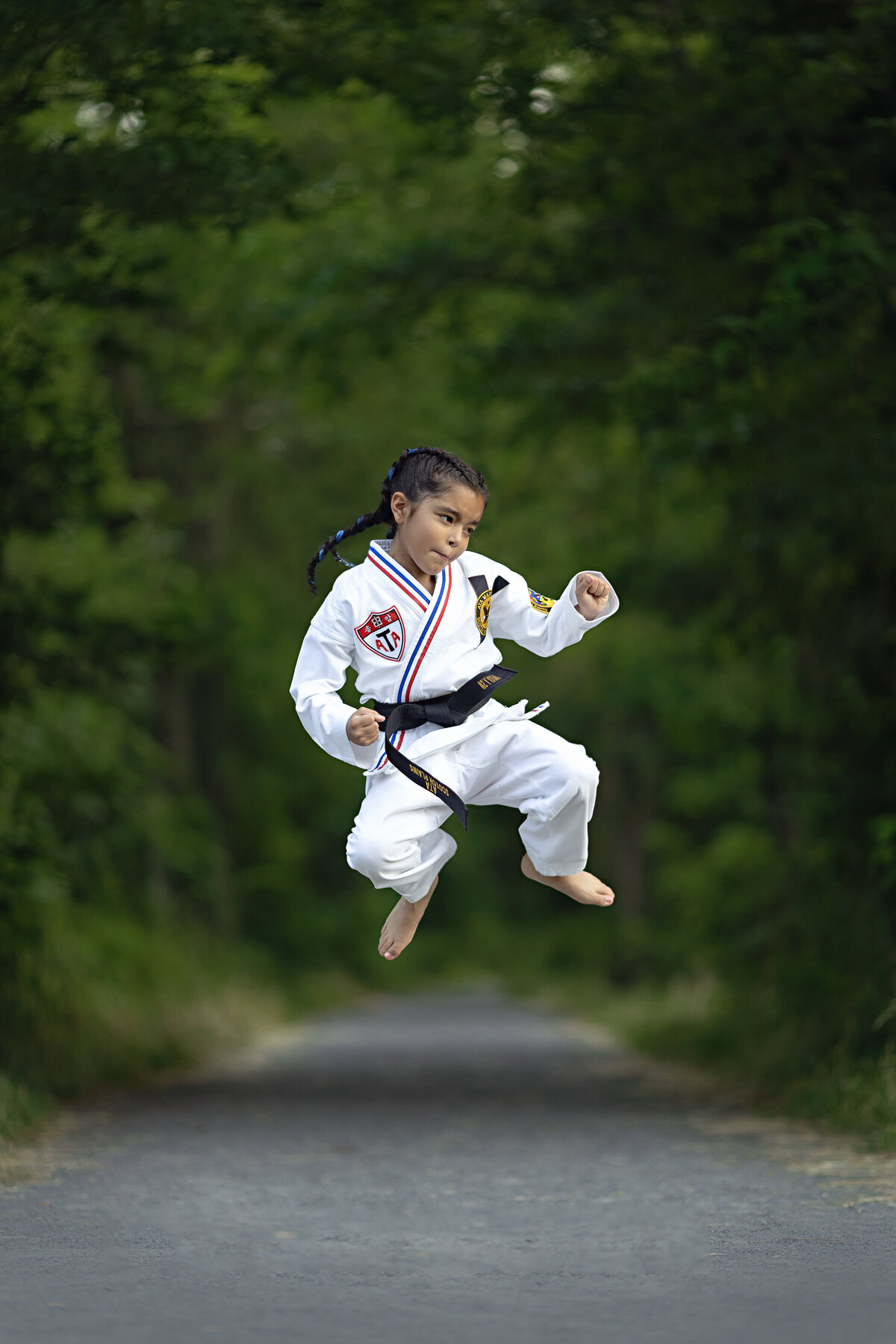 A young girl in a karate uniform performs a jump kick in a park sidewalk