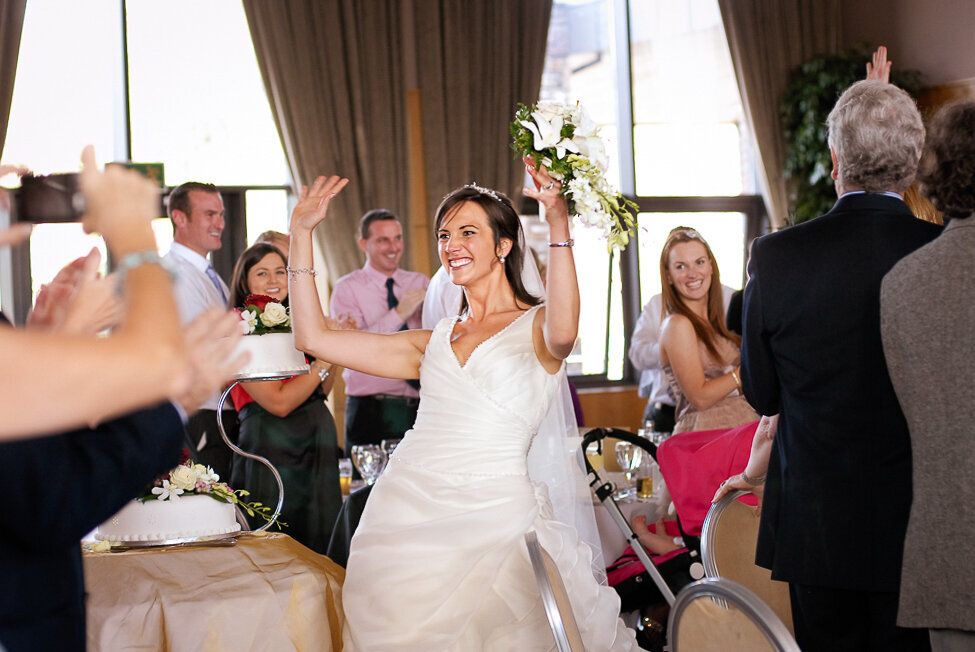 bride wearing a princees style wedding dress with tulle dancing her way into the wedding reception, holding white bouquet in the air while her wedding guests cheer