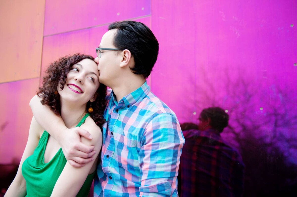 a man kisses a womans head in front of the bright pink wall of the pop museum in seattle